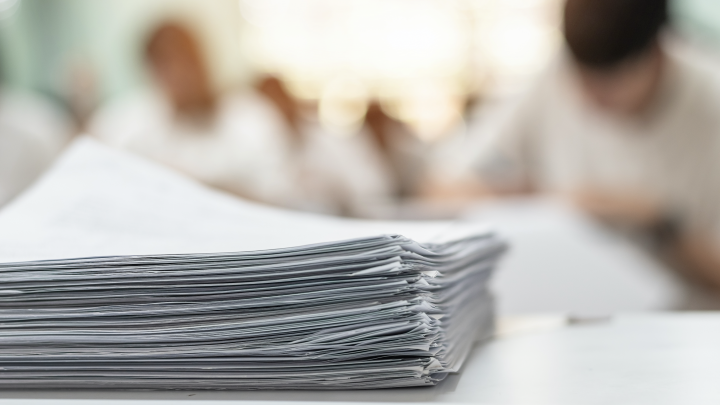 Photo of a stack of papers in focus and several students in the background who are out of focus.