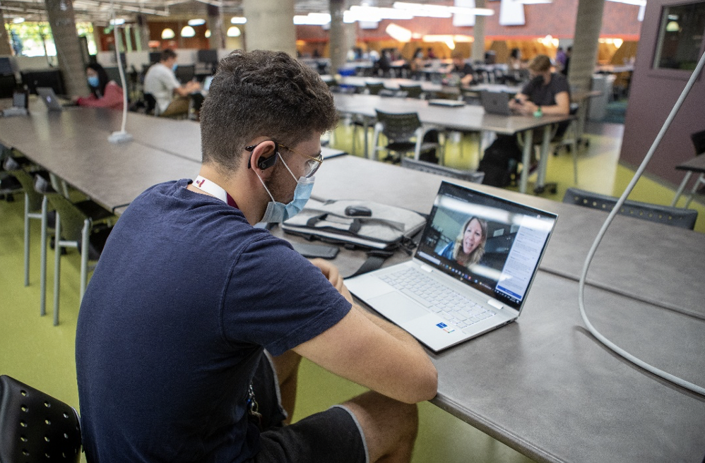 A person in a university library wearing a mask and sitting at a table with a computer. He is watching an online lecture.